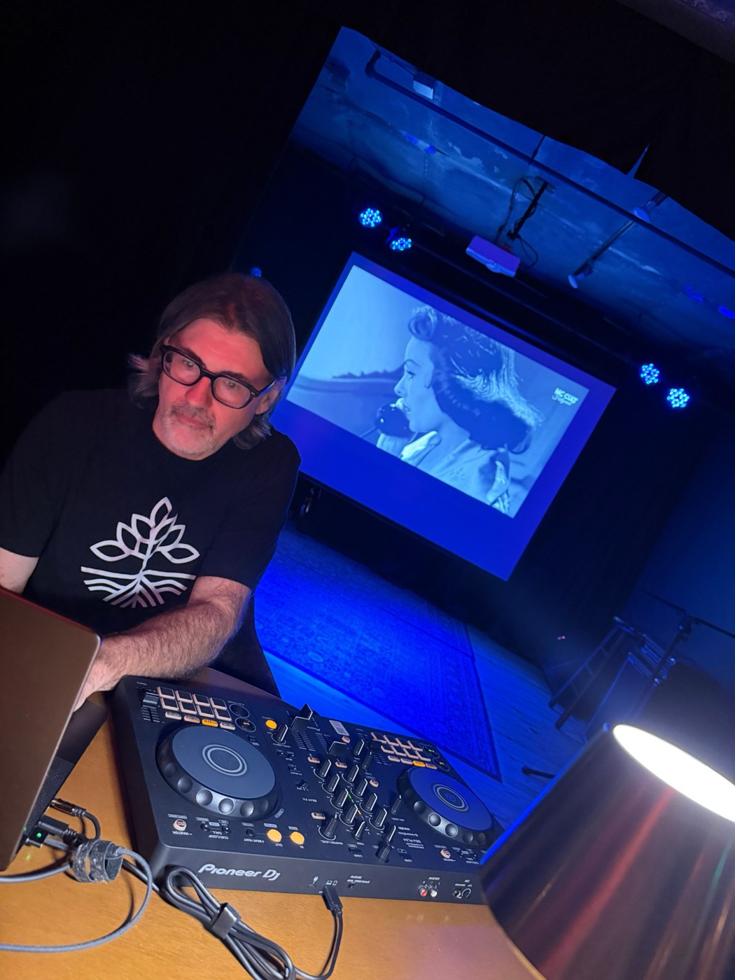 A DJ at the decks with an old film playing on the screen behind